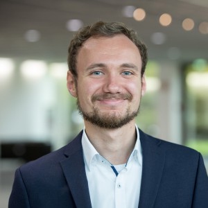 Das Foto zeigt einen Mann mit blauem Sakko. Er hat kurze, lockige Haare und einen Bart. Er trägtein weißes Hemd ohne Krawatte