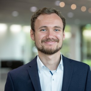 Das Foto zeigt einen Mann mit blauem Sakko. Er hat kurze, lockige Haare und einen Bart. Er trägtein weißes Hemd ohne Krawatte.