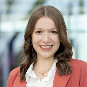Das Foto zeigt eine Fraut mit brunetten Haaren. Sie trägt einen roten Blazer über einem weißen Hemd. Sie lächelt in die Kamera.
