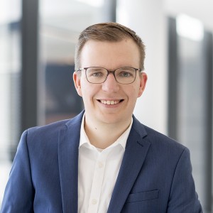 Das Foto zeigt einen Mann mit blauem Sakko. Er hat kurze Haare und eine Brille. Er trägt Jeans, ein weißes Hemd ohne Krawatte und lehnt an ein Geländer.