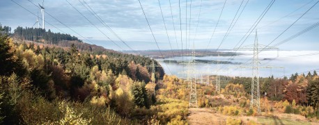 Das Foto zeigt eine Trasse in einer bewaldeten Hügellandschaft. Im Hintergrund sind zwei Windräder zu sehen.