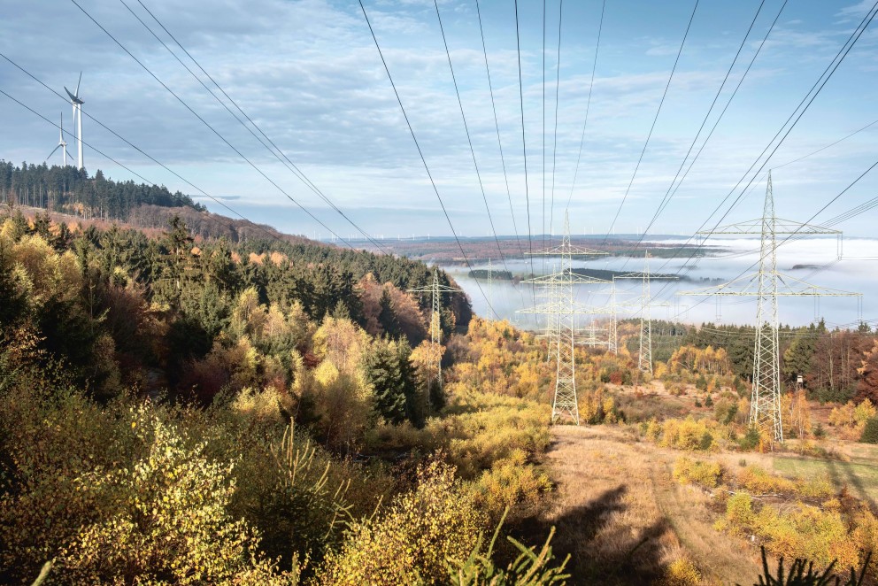 Das Foto zeigt eine Trasse in einer bewaldeten Hügellandschaft. Im Hintergrund sind zwei Windräder zu sehen.